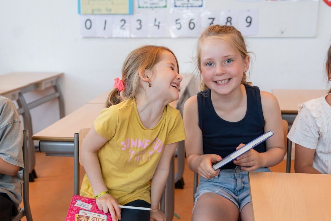 Twee lachende meisjes in de klas, een met een schrift in haar handen